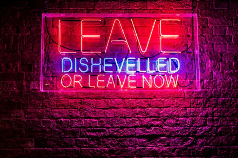 Red and blue neon on a birkc wass reads "Leave dishevelled or leave now."